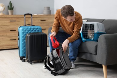 Photo of Travel with pet. Man putting headphones into backpack near carrier with cute cat at home