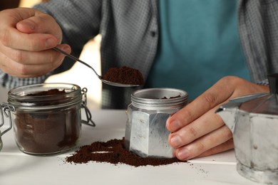 Man putting ground coffee into moka pot at white wooden table indoors, closeup