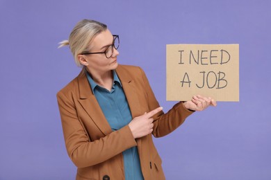 Unemployed senior woman pointing at cardboard sign with phrase I Need A Job on purple background