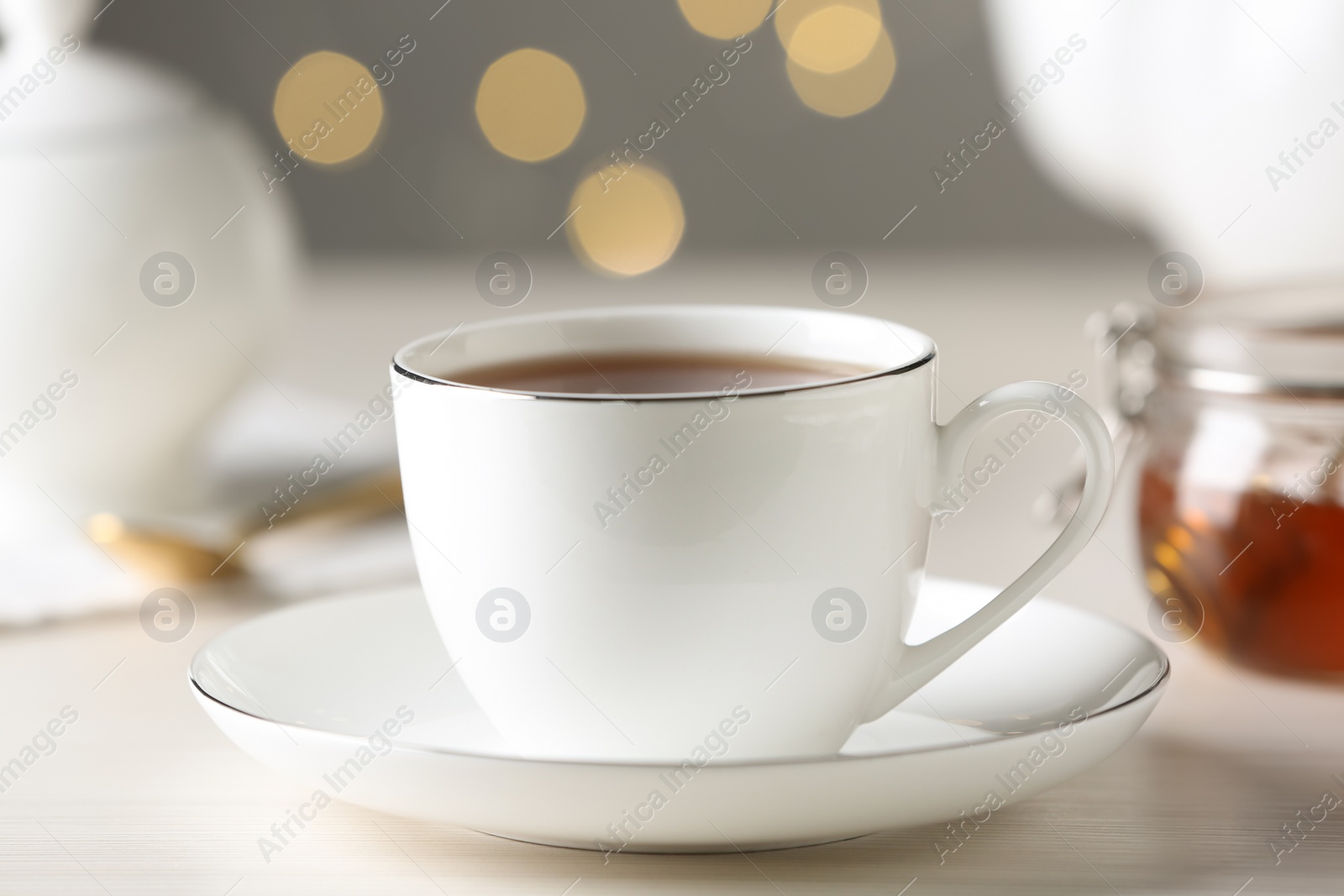 Photo of Cup of hot tea with saucer on white table against blurred lights, closeup