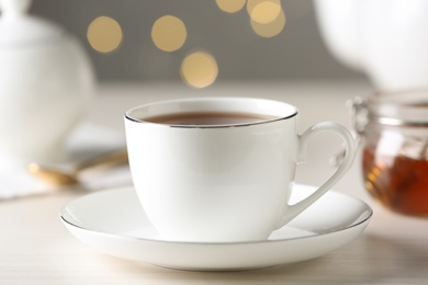Cup of hot tea with saucer on white table against blurred lights, closeup