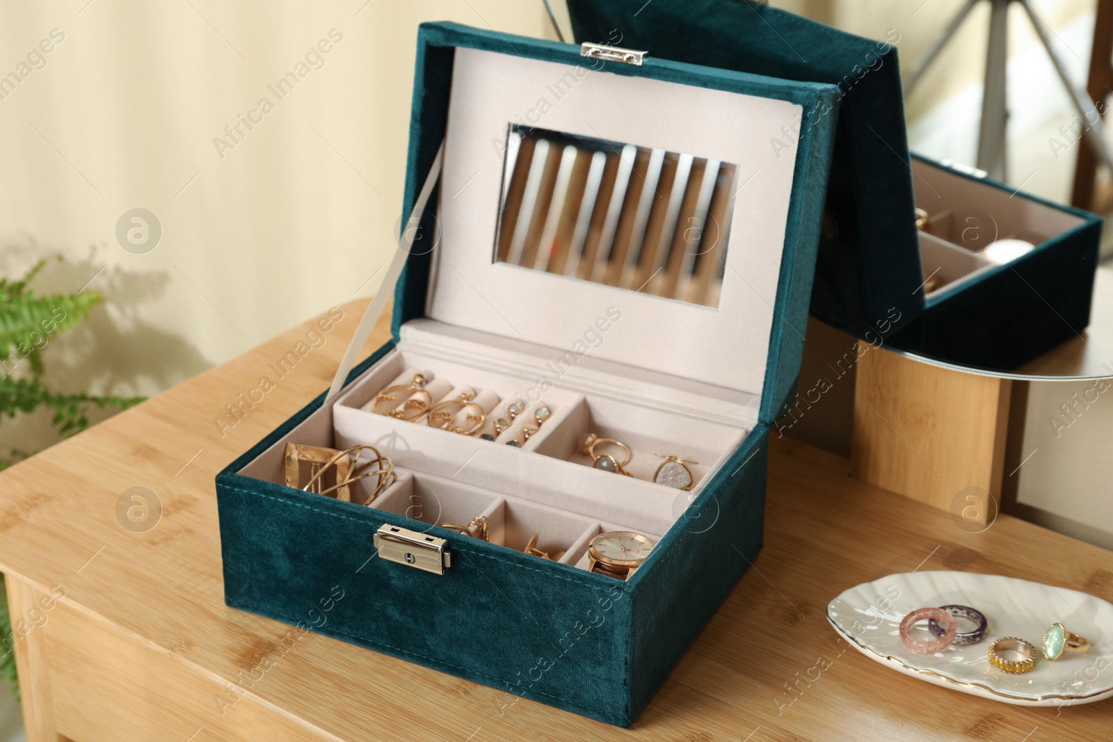 Photo of Elegant jewelry box with beautiful bijouterie and wristwatch near mirror on wooden table in room