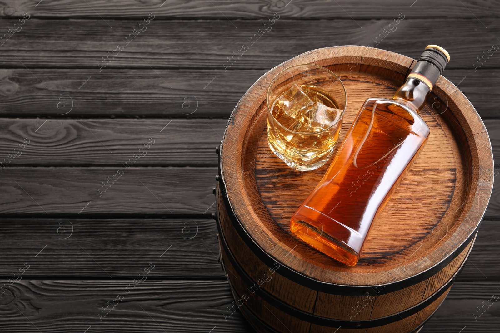 Photo of Whiskey with ice cubes in glass, bottle and barrel on black wooden table, above view. Space for text