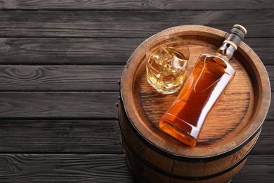 Photo of Whiskey with ice cubes in glass, bottle and barrel on black wooden table, above view. Space for text
