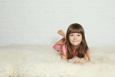 Photo of Cute little girl lying on fur rug against brick wall. Space for text