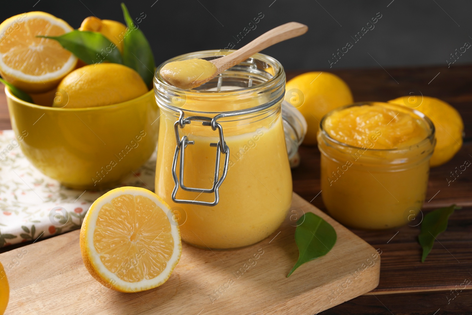 Photo of Delicious lemon curd in glass jars, spoon, fresh citrus fruits and green leaves on wooden table