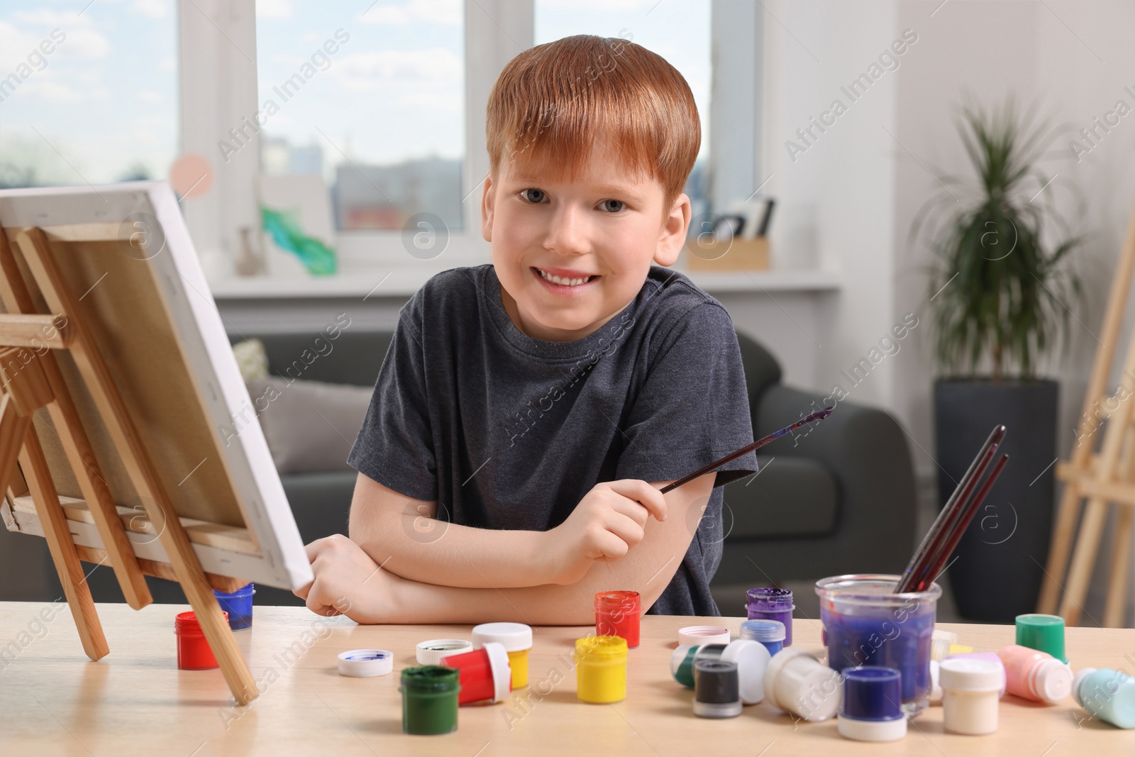 Photo of Happy little boy painting at table in studio. Using easel to hold canvas