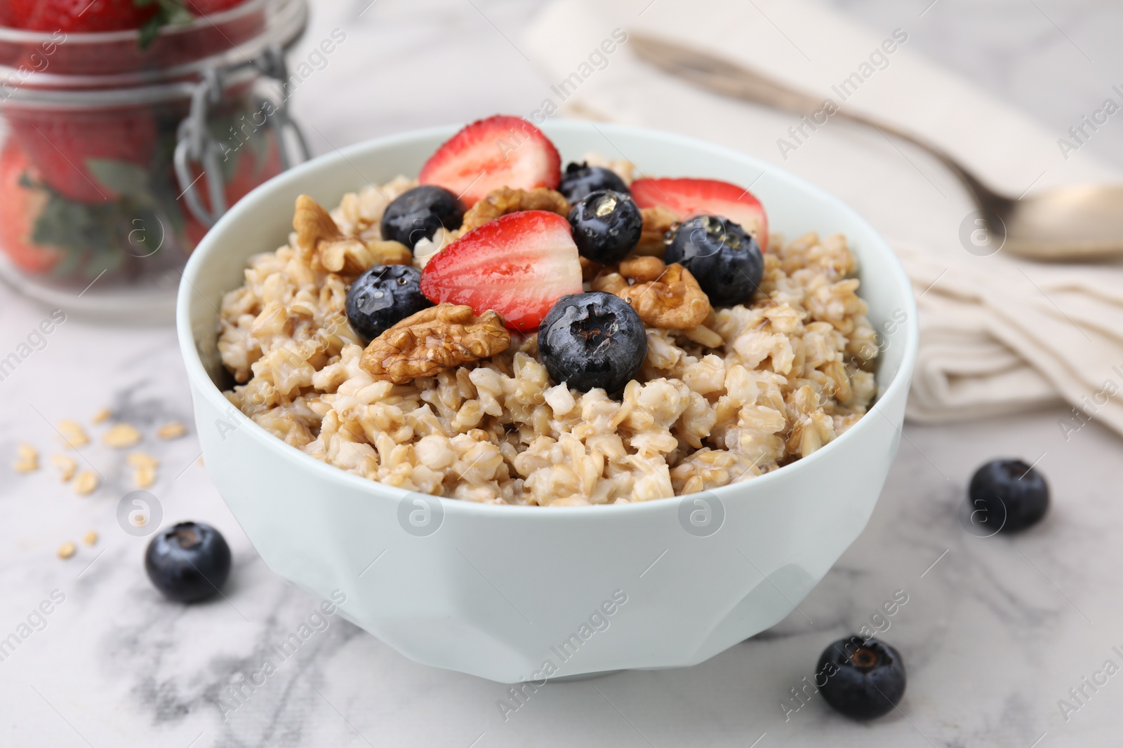 Photo of Tasty oatmeal with strawberries, blueberries and walnuts in bowl on white marble table, closeup