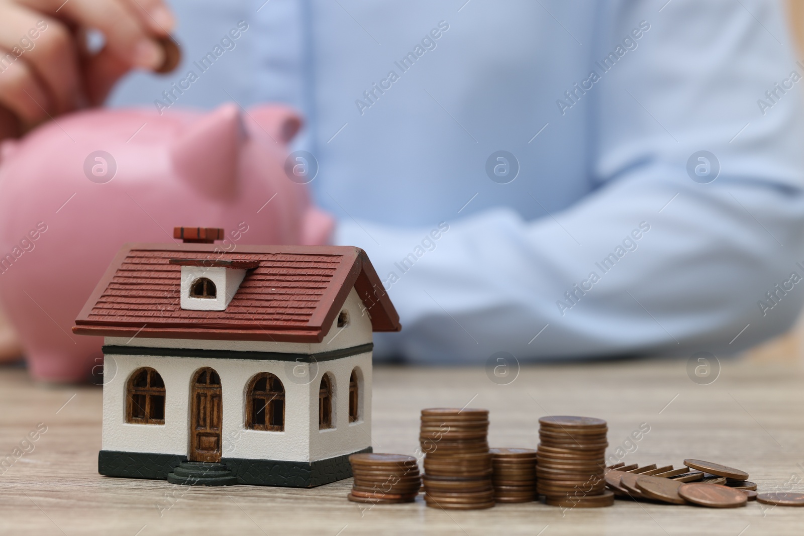 Photo of Woman with piggy bank at wooden table, focus on house model and stacked coins