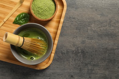 Cup of fresh matcha tea with bamboo whisk, green powder and spoon on dark grey table, top view. Space for text