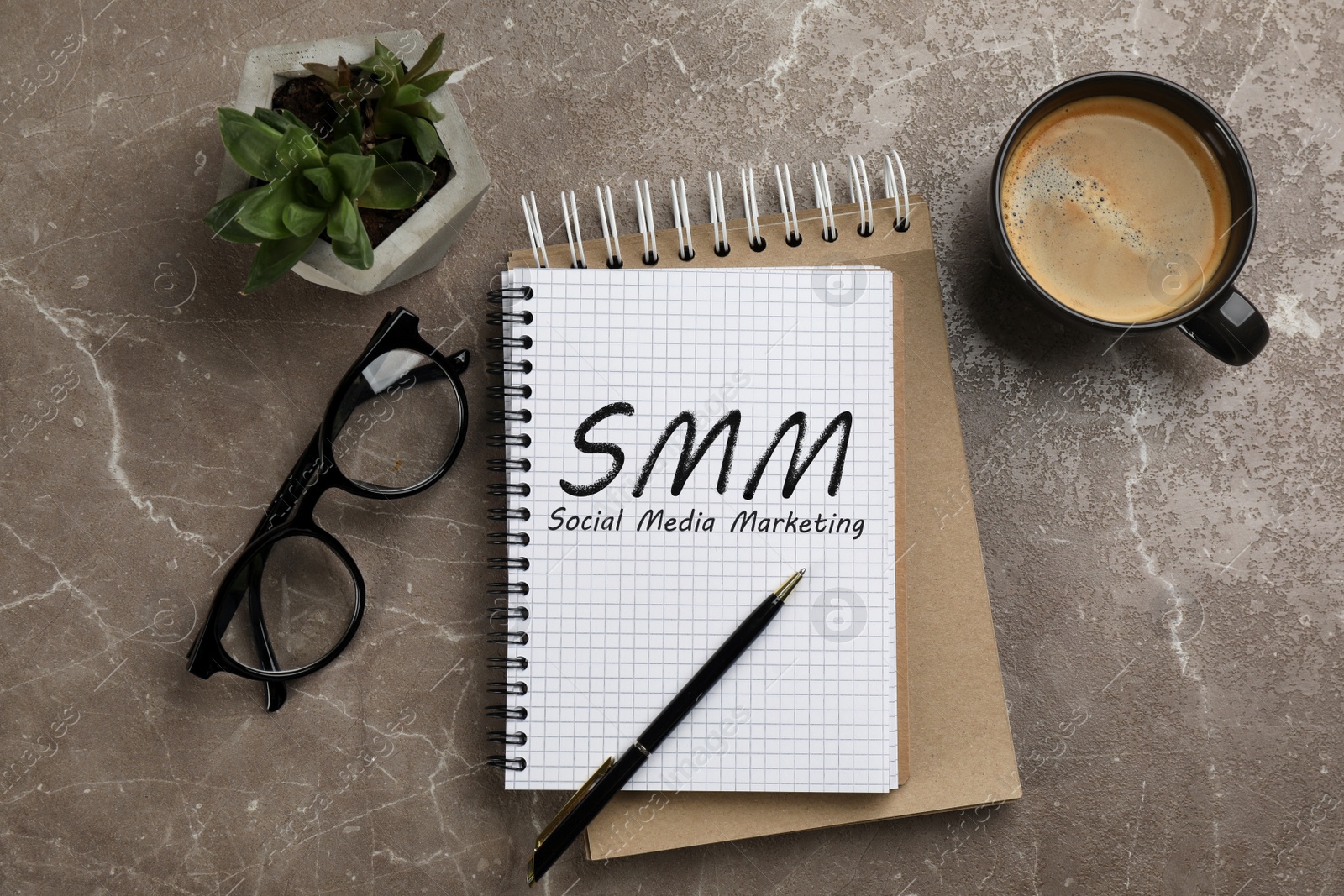 Image of Notebook with text SMM (Social media marketing), cup of aromatic coffee, glasses and plant on brown marble table, flat lay