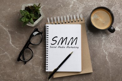 Notebook with text SMM (Social media marketing), cup of aromatic coffee, glasses and plant on brown marble table, flat lay
