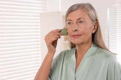 Woman massaging her face with jade gua sha tool in bathroom. Space for text