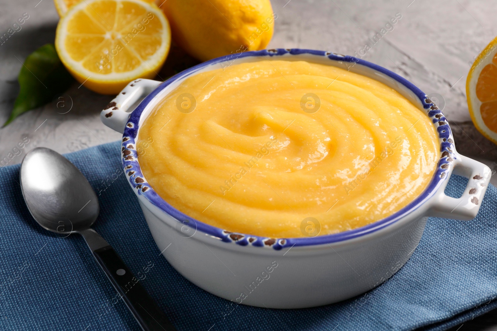 Photo of Delicious lemon curd in bowl, fresh citrus fruits and spoon on grey table, closeup