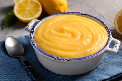 Photo of Delicious lemon curd in bowl, fresh citrus fruits and spoon on grey table, closeup