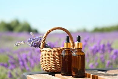 Photo of Bottles of essential oil and wicker bag with lavender flowers on wooden table in field outdoors, space for text