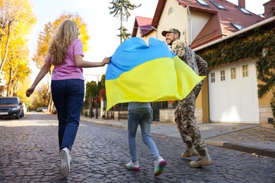 Photo of Soldier in military uniform with his family running and holding Ukrainian flag on city street, back view