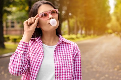Photo of Beautiful young woman with pink sunglasses blowing chewing gum outdoors, space for text