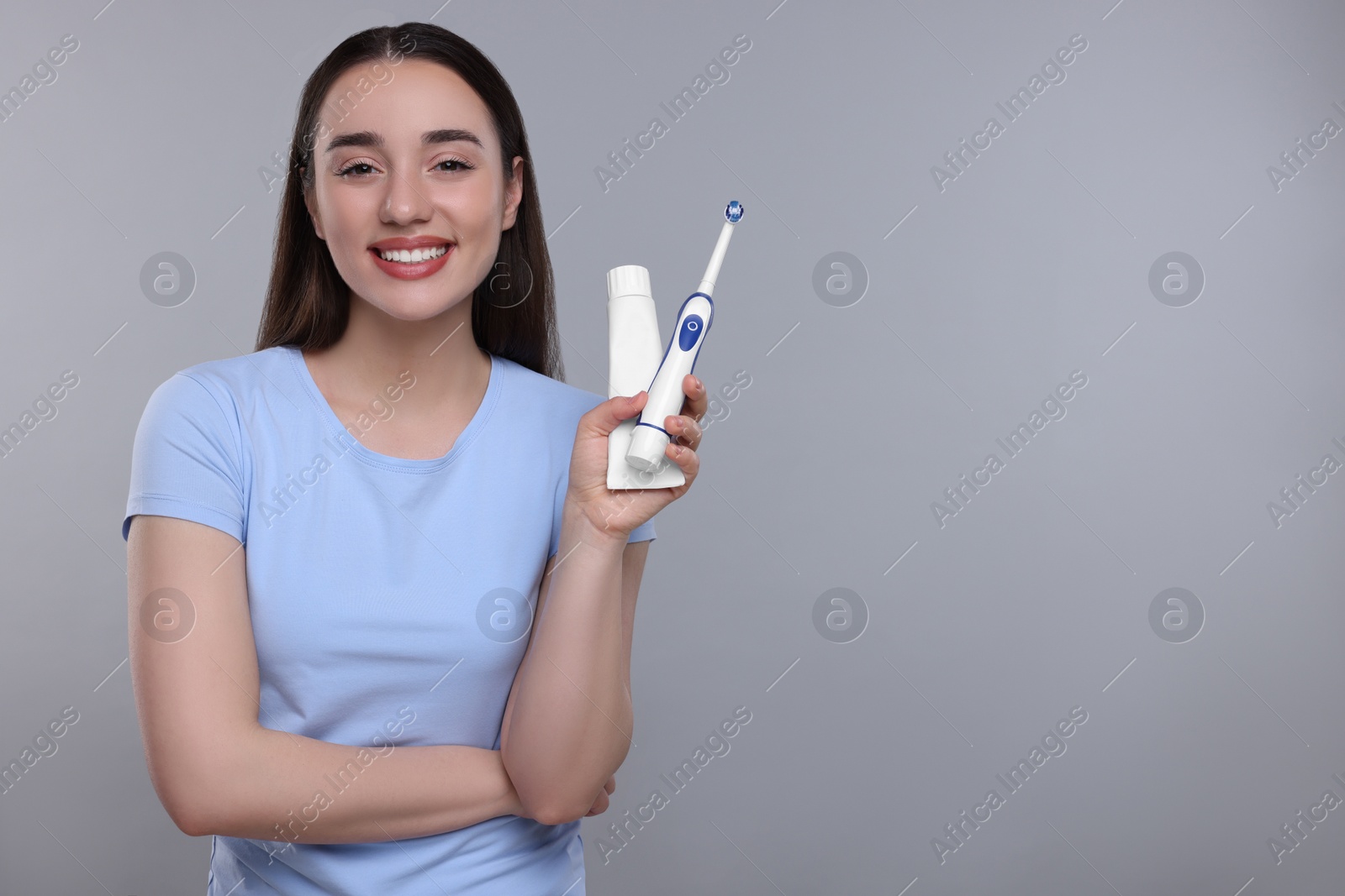 Photo of Happy young woman holding electric toothbrush and tube of toothpaste on light grey background, space for text
