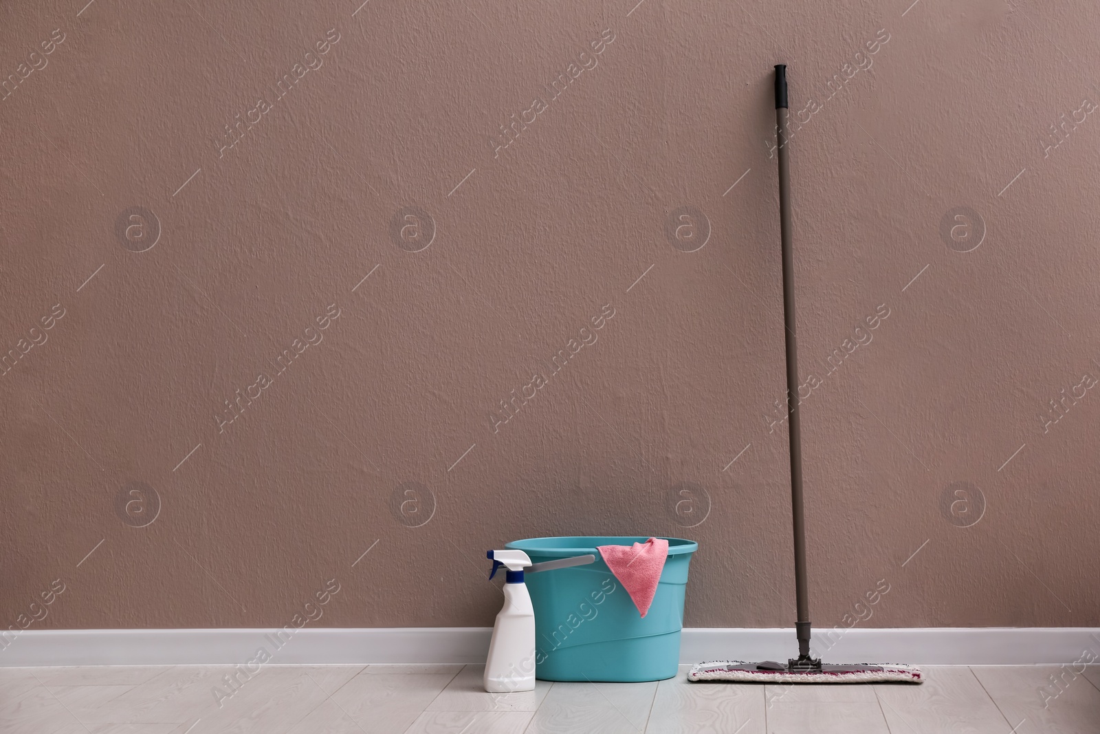 Photo of Floor mop, cleaning detergent and bucket with rag near brown wall indoors. Space for text