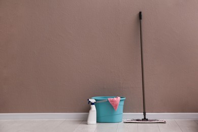 Photo of Floor mop, cleaning detergent and bucket with rag near brown wall indoors. Space for text