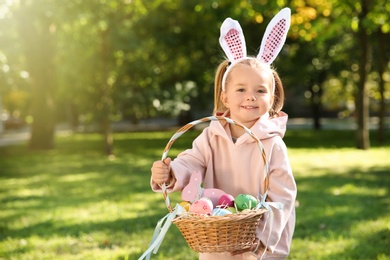 Photo of Cute little girl with bunny ears and basket of Easter eggs in park. Space for text