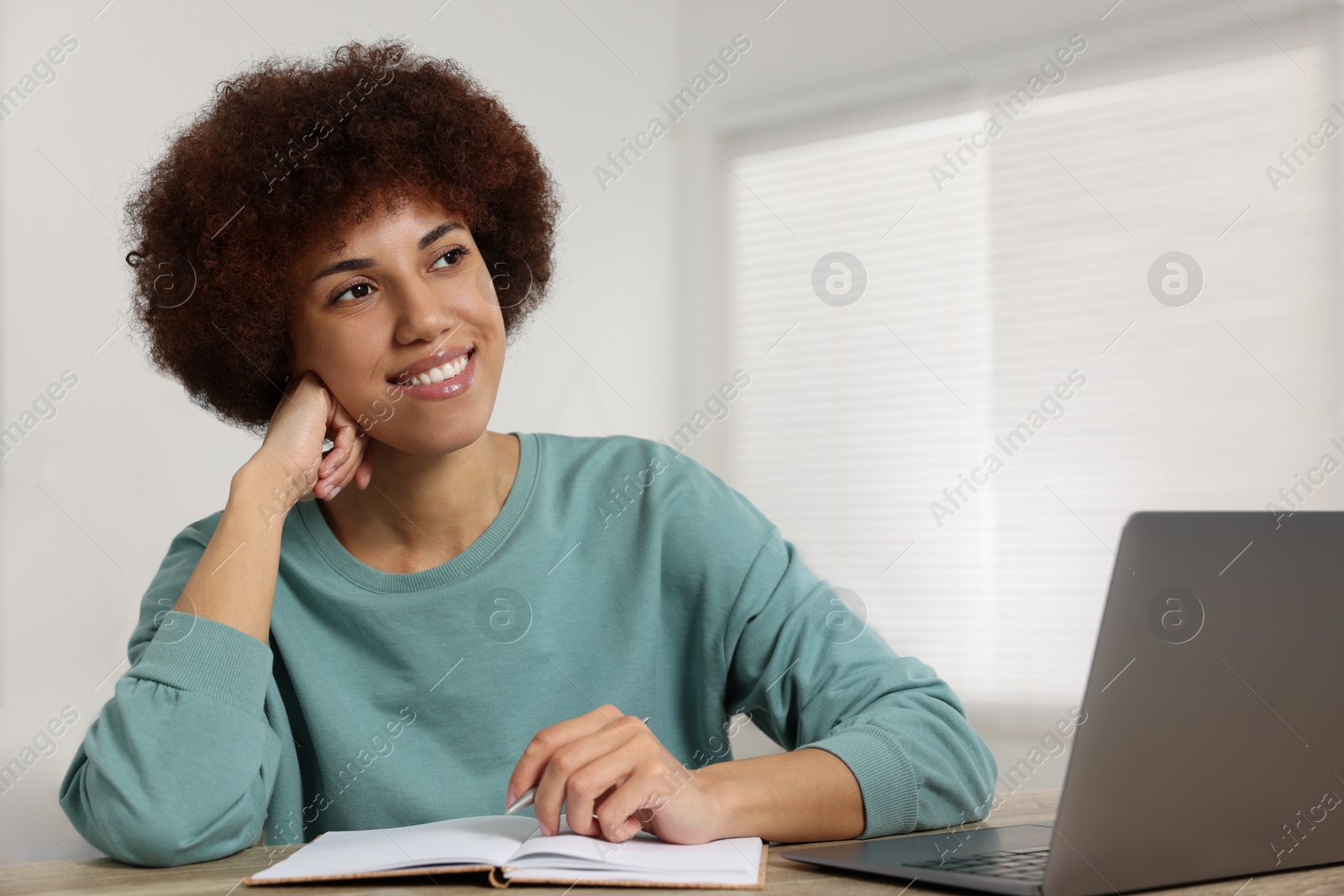Photo of Young woman using laptop and writing in notebook at wooden desk in room