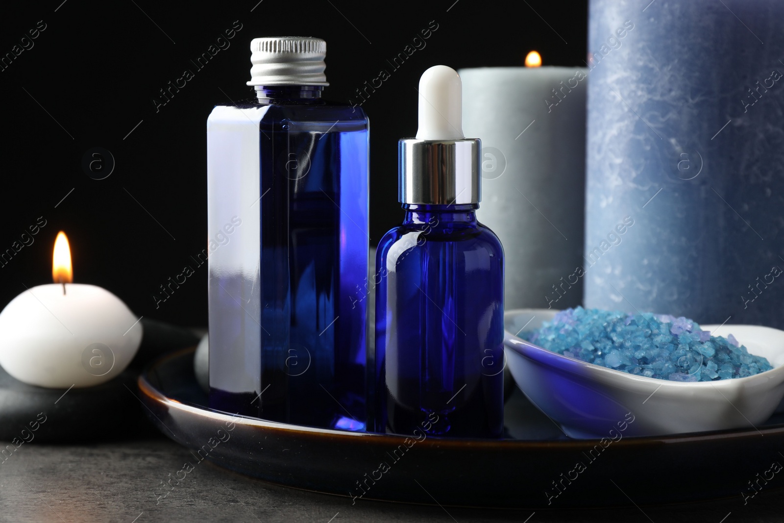 Photo of Spa composition. Cosmetic products, burning candles and sea salt on gray table against black background, closeup