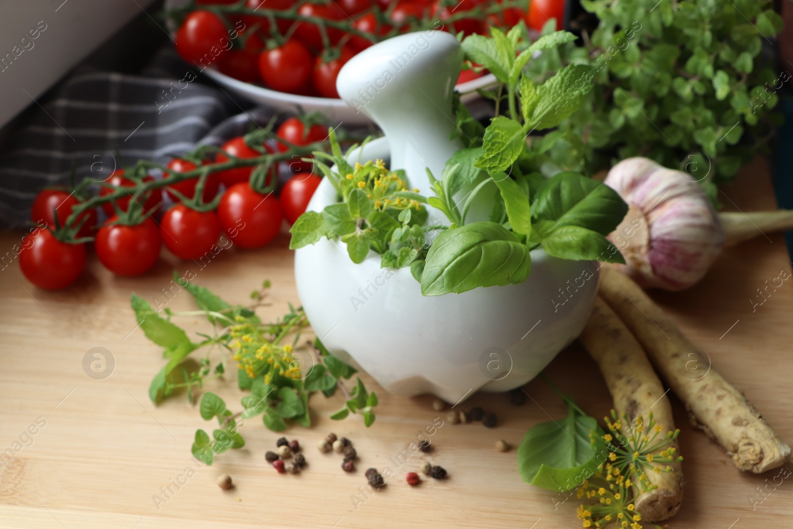 Photo of Mortar with fresh herbs near garlic, horseradish roots, black peppercorns and cherry tomatoes on wooden table, closeup