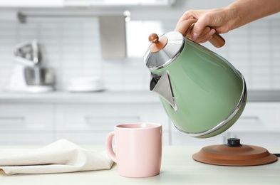 Photo of Woman pouring water from modern electric kettle into cup at wooden table in kitchen, closeup