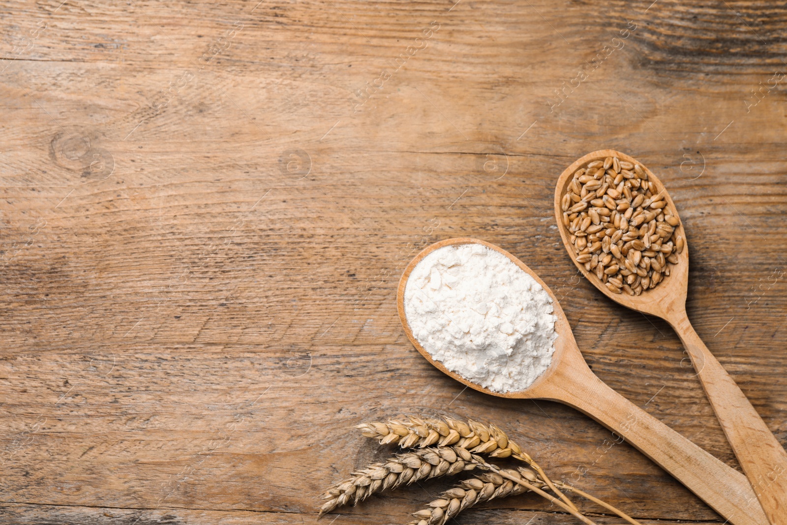 Photo of Spoons with wheat flour, grains and spikes on wooden table, flat lay. Space for text