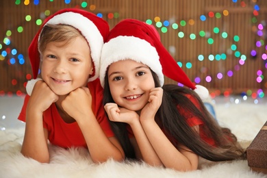 Photo of Cute little children in Santa hats lying on floor against blurred lights. Christmas celebration
