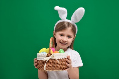 Easter celebration. Cute girl with bunny ears holding basket of painted eggs on green background