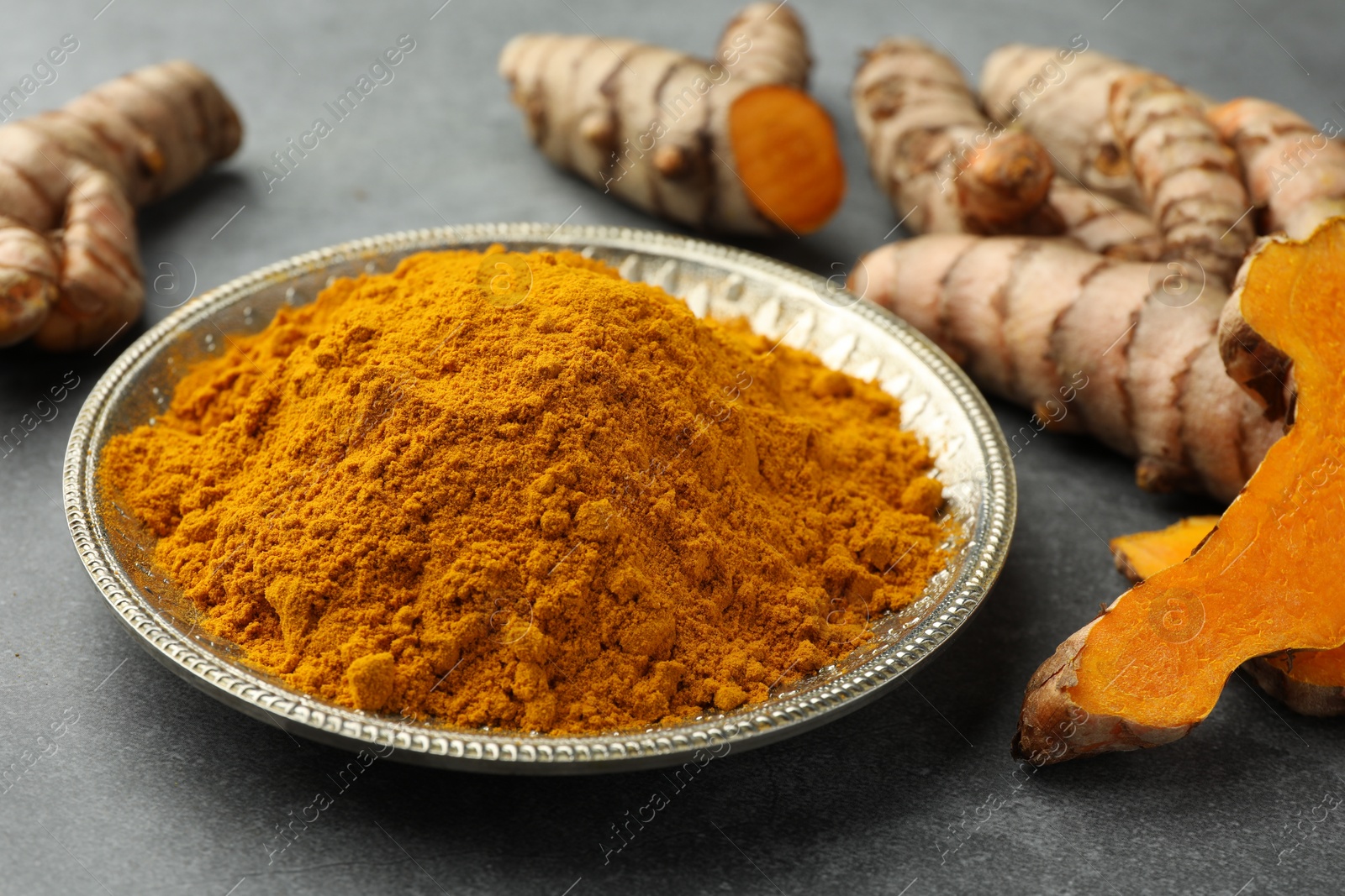 Photo of Plate with turmeric powder and raw roots on grey table, closeup