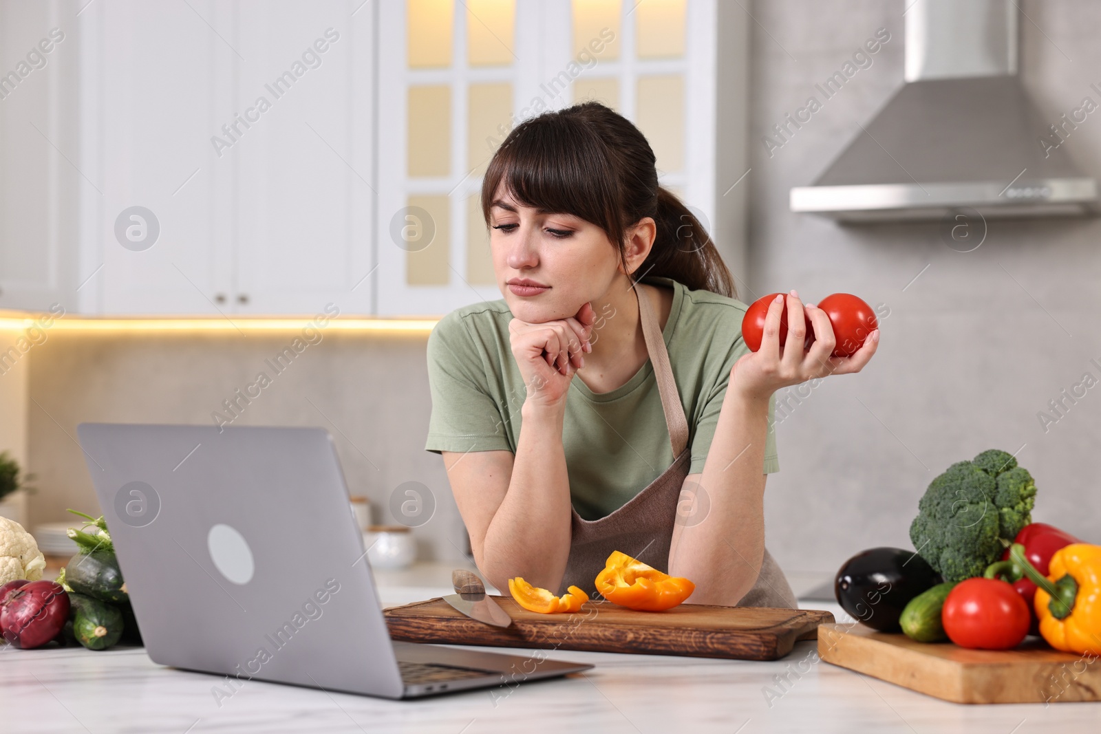 Photo of Young housewife cooking while watching online course via laptop at white marble table in kitchen