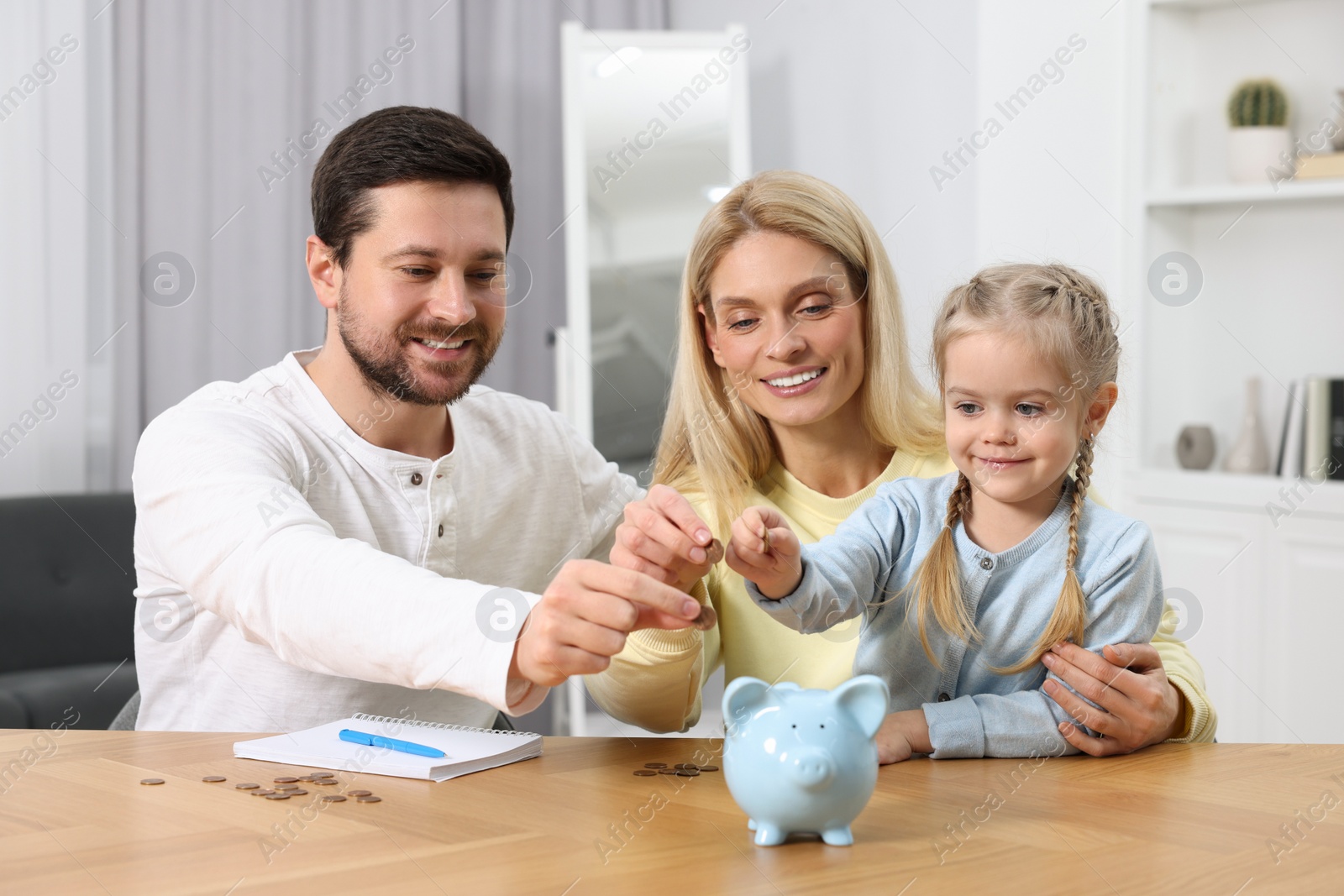 Photo of Planning budget together. Little girl with her family putting coins into piggybank at table indoors