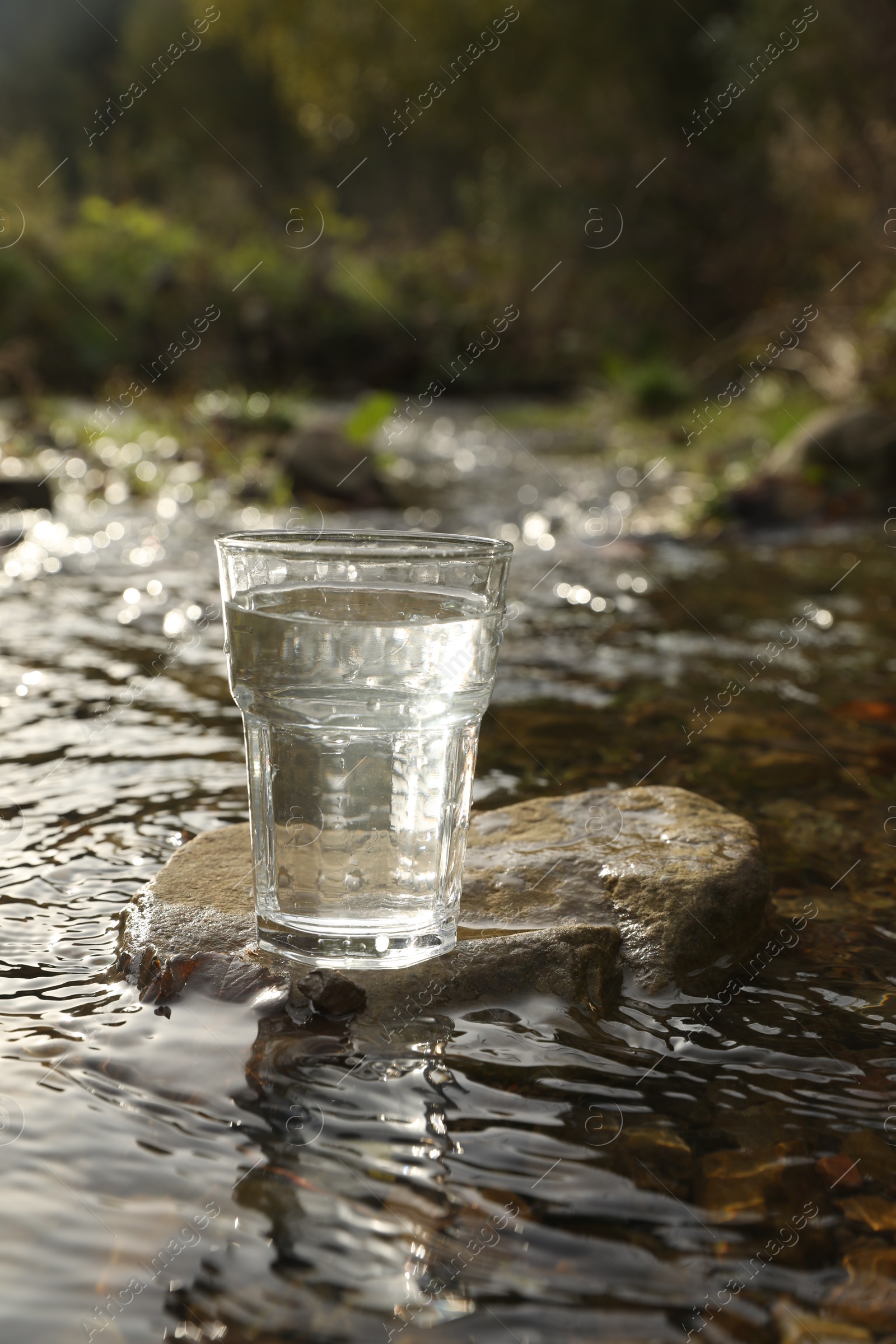 Photo of Glass of fresh water on stone near river outdoors. Space for text