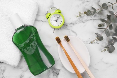 Fresh mouthwash in bottle, toothbrushes and alarm clock on white marble table, flat lay