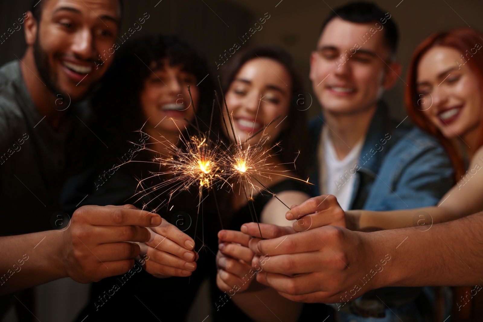 Photo of Happy friends with sparklers celebrating birthday indoors, focus on hands