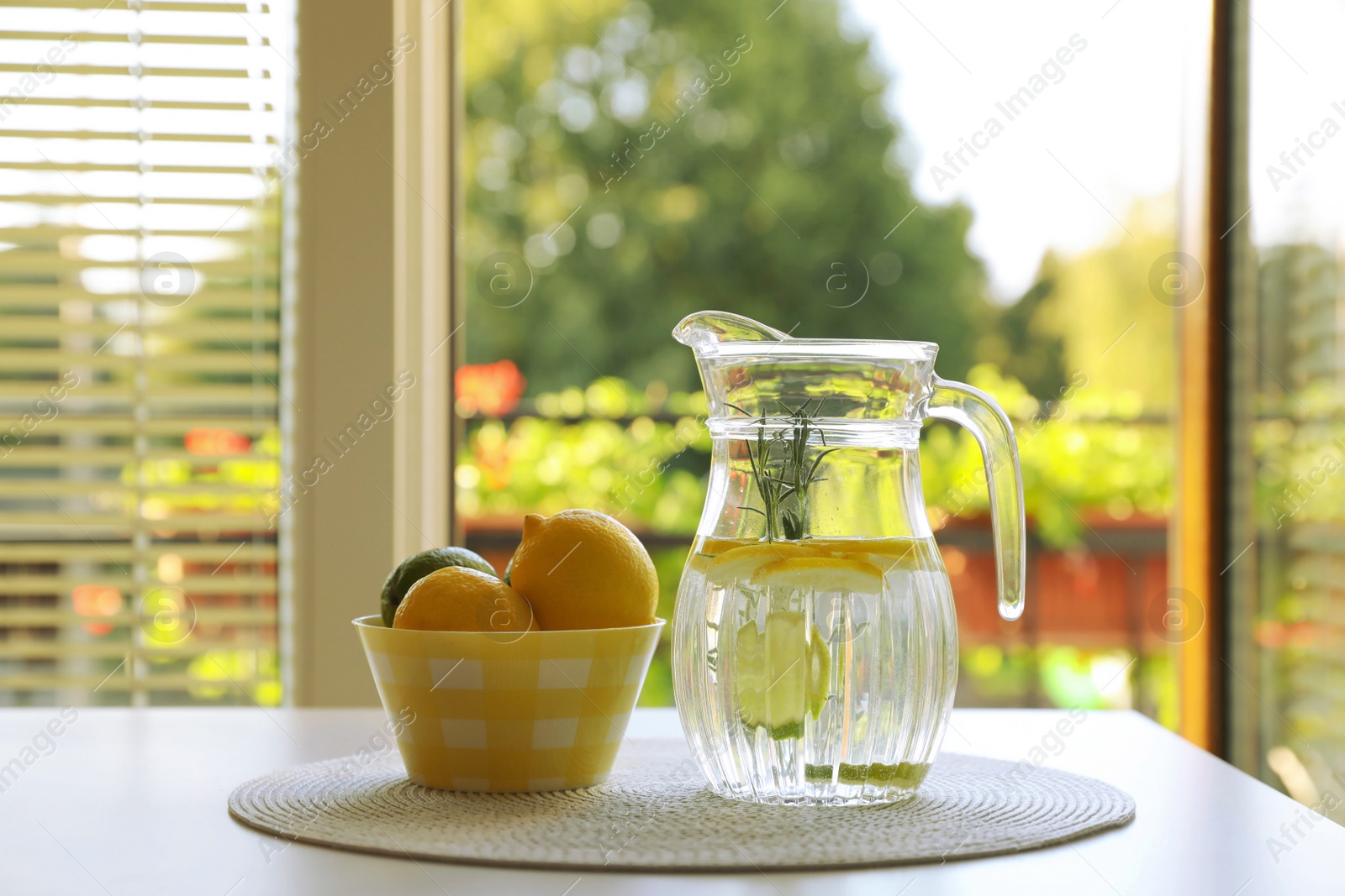 Photo of Jug with refreshing lemon water and citrus fruits in bowl on table indoors