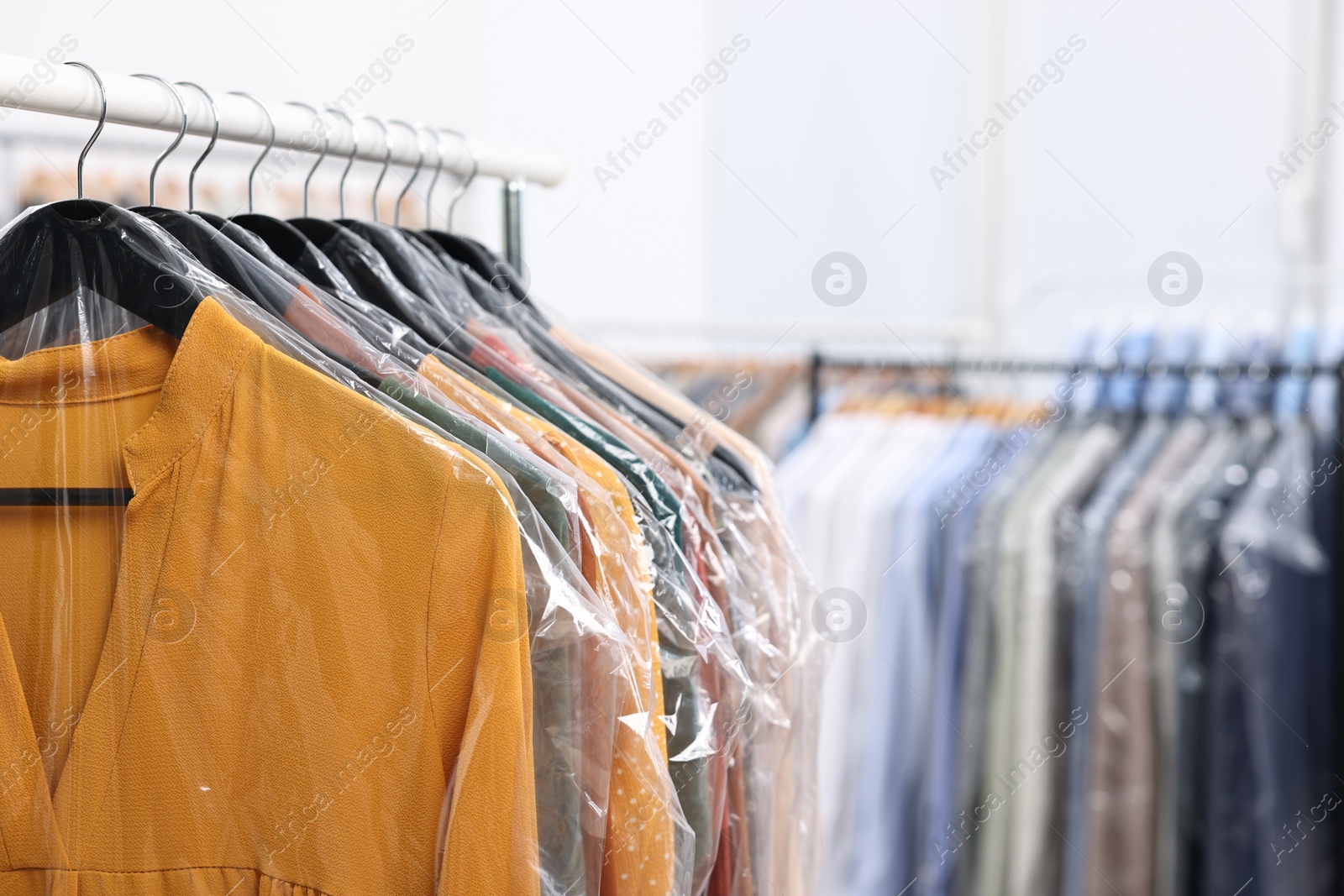 Photo of Dry-cleaning service. Many different clothes in plastic bags hanging on rack indoors, closeup