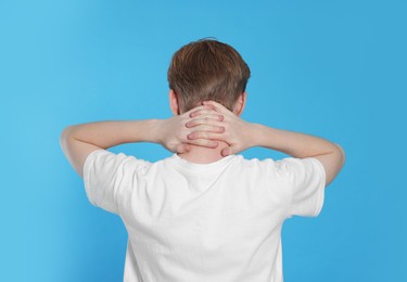 Photo of Teenage boy suffering from pain in neck on light blue background, back view. Arthritis symptom