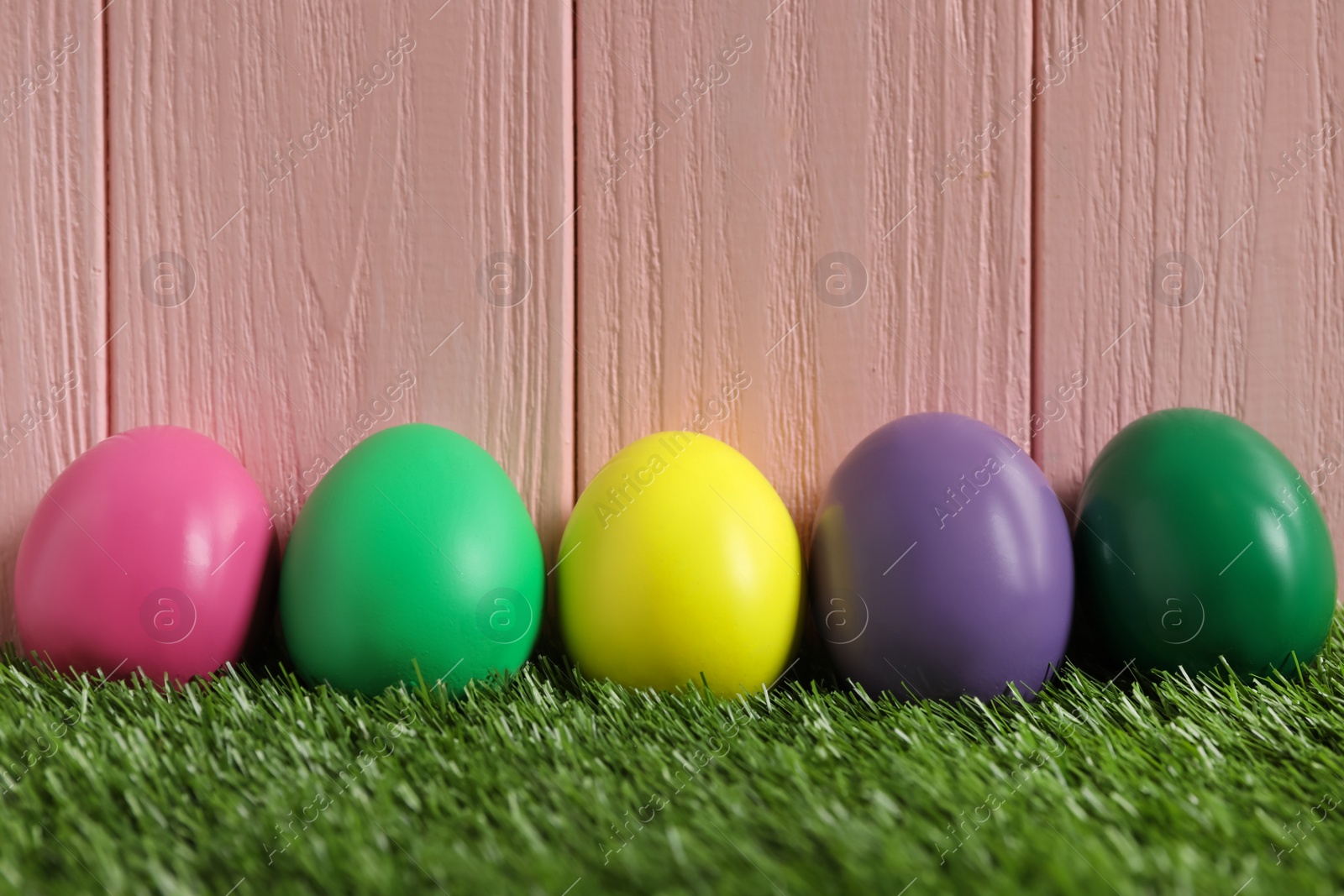 Photo of Bright Easter eggs on green grass against pink wooden background