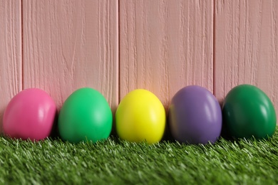 Photo of Bright Easter eggs on green grass against pink wooden background