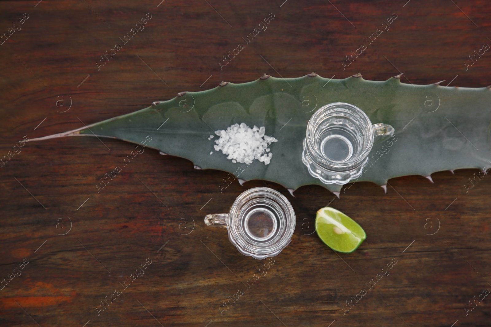 Photo of Mexican tequila shots, salt, lime slice and green leaf on wooden table, flat lay. Drink made of agava