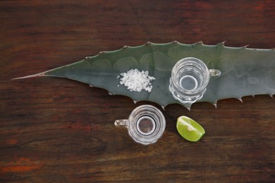 Photo of Mexican tequila shots, salt, lime slice and green leaf on wooden table, flat lay. Drink made of agava