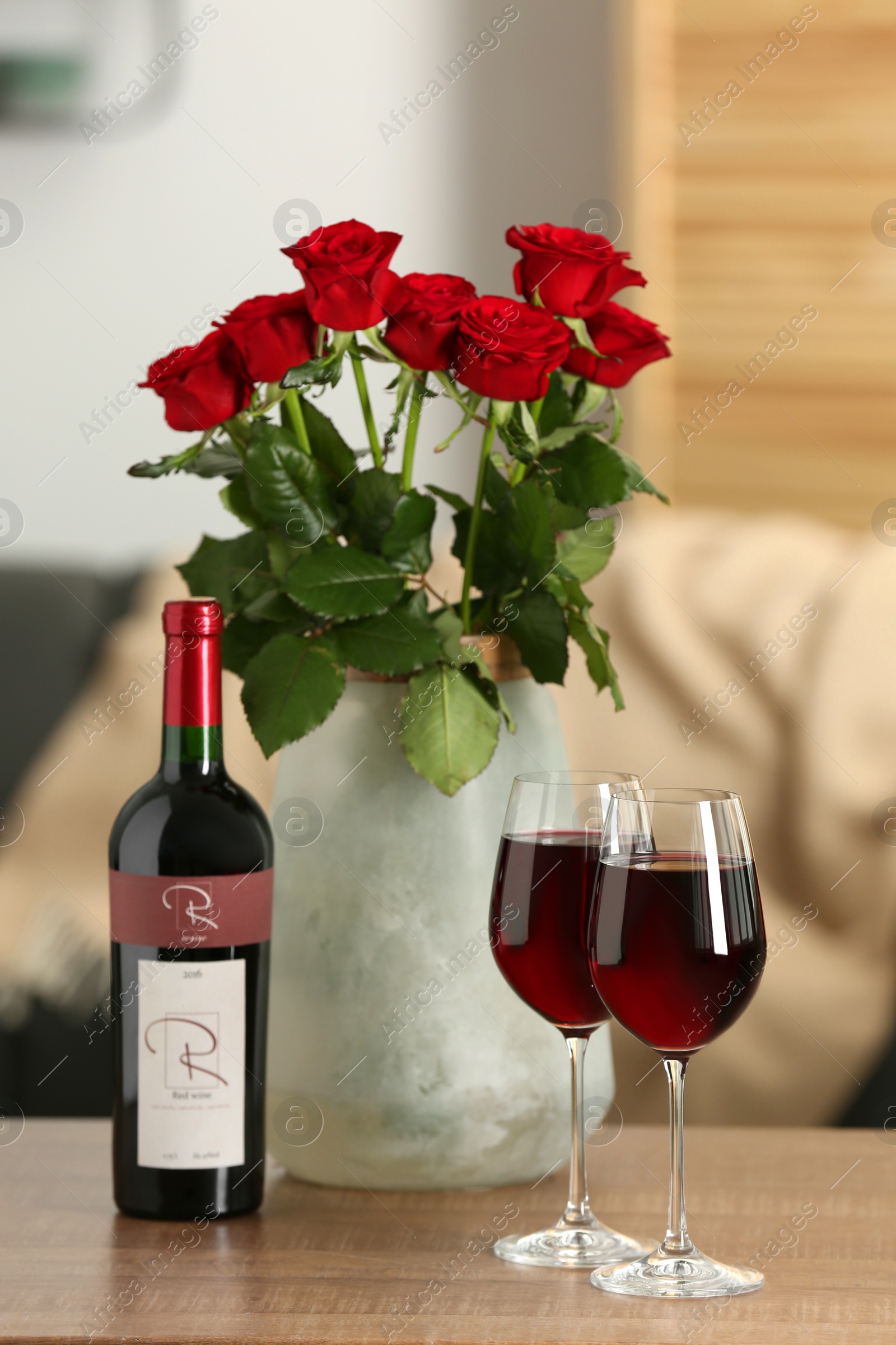 Photo of Bottle, glasses of red wine and vase with roses on wooden table in room. Romantic date