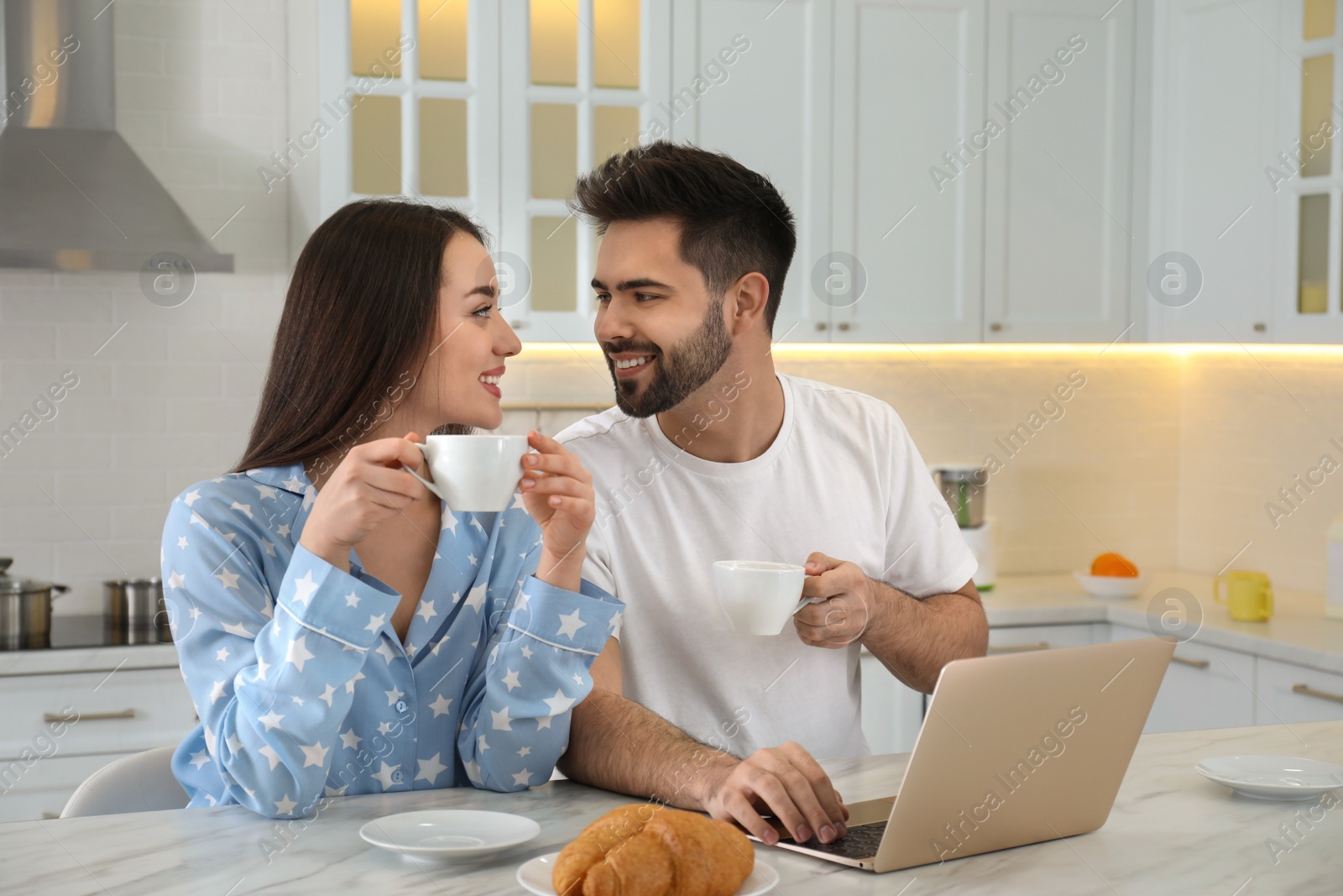 Photo of Happy couple in pajamas with laptop having breakfast at kitchen table