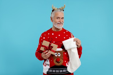 Photo of Senior man in Christmas sweater, reindeer headband holding gift and stocking on light blue background