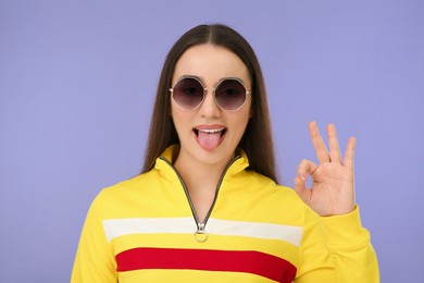 Happy woman in sunglasses showing her tongue and OK gesture on violet background